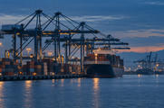 Synthetic editorial illustration. Cargo vessel, containers, and gantry cranes at an unbranded port at blue hour.