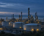 Synthetic editorial illustration. Unbranded refinery storage tanks and distillation columns at blue-hour dawn for energy-market news.