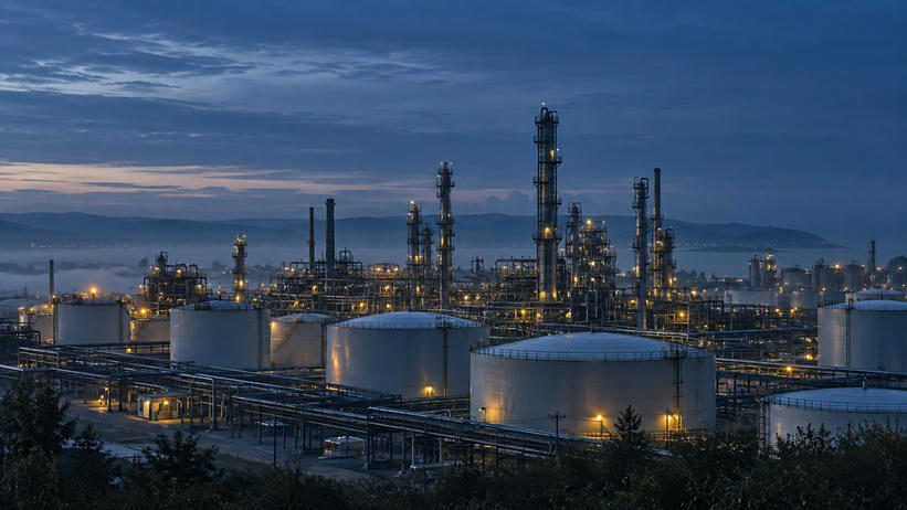 Synthetic editorial illustration. Unbranded refinery storage tanks and distillation columns at blue-hour dawn for energy-market news.