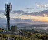 Synthetic editorial illustration. Unbranded cellular relay mast above a misty rural landscape for telecom and connectivity news.