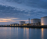 Synthetic editorial illustration. Unbranded waterfront energy terminal with storage tanks and pipework for energy-market news.