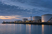 Synthetic editorial illustration. Unbranded waterfront energy terminal with storage tanks and pipework for energy-market news.