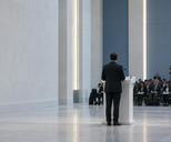 Synthetic editorial illustration. Anonymous speaker viewed from behind at a blank lectern in an unbranded policy briefing room.