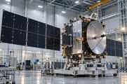 Synthetic editorial illustration. Unbranded communications satellite being assembled in a high-bay aerospace integration facility.