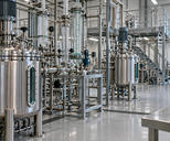 Synthetic editorial illustration. Unbranded stainless process vessels and glass pipework in a specialty-chemicals pilot plant.