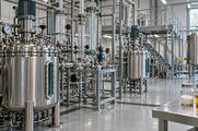 Synthetic editorial illustration. Unbranded stainless process vessels and glass pipework in a specialty-chemicals pilot plant.