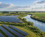 Synthetic editorial illustration. Solar panels and distant wind turbines beside a curving waterway in a lowland agricultural landscape.