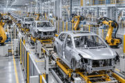 Synthetic editorial illustration. Unbranded electric vehicle bodies and robotic arms on a modern automotive assembly line.