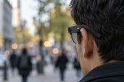 Synthetic editorial illustration. A city street. A tight rear-side view shows an ear and dark hair beneath a plain glasses frame with a small lens and glowing indicator; pedestrians and buildings are softly blurred behind.
