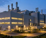 Synthetic editorial illustration. A semiconductor factory exterior at dusk. A large grey building has brightly lit window bands, rooftop exhaust stacks and utility pipework beside quiet access roads and landscaping.