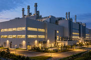 Synthetic editorial illustration. A semiconductor factory exterior at dusk. A large grey building has brightly lit window bands, rooftop exhaust stacks and utility pipework beside quiet access roads and landscaping.