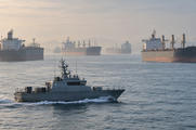 Synthetic editorial illustration. An offshore anchorage. Several large cargo ships wait on hazy water while a small grey patrol boat crosses the foreground, trailing a low white wake beneath muted light.