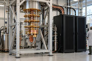 Synthetic editorial illustration. A tiered gold-and-copper cryostat hangs in a steel frame beside two black server cabinets linked by cables. A grey-haired engineer viewed from behind stands in the daylit laboratory.