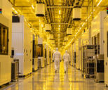 Synthetic editorial illustration. Two white-suited technicians walk away down a reflective cleanroom aisle under yellow lighting. Plain equipment enclosures flank the aisle, with suspended carrier pods and transport rails overhead.