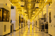 Synthetic editorial illustration. Two white-suited technicians walk away down a reflective cleanroom aisle under yellow lighting. Plain equipment enclosures flank the aisle, with suspended carrier pods and transport rails overhead.