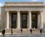 Synthetic editorial illustration. A neoclassical institutional facade has four tall columns and broad stone steps. Small rear-facing pedestrians approach its dark glazed entrances in daylight.