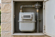 Synthetic editorial illustration. One plain household gas meter with blank counter window and simple gas pipework in an open wall cabinet.