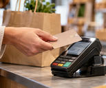 Synthetic editorial illustration. An adult hand holds a plain card over a powered checkout terminal beside a filled paper shopping bag in a softly defocused shop.