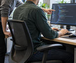 Synthetic editorial illustration. Two anonymous colleagues working together at a desk with a complete illuminated monitor directly in front of the seated engineer, who is using a keyboard; the colleague gestures toward softly rendered editor-like content.
