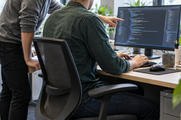 Synthetic editorial illustration. Two anonymous colleagues working together at a desk with a complete illuminated monitor directly in front of the seated engineer, who is using a keyboard; the colleague gestures toward softly rendered editor-like content.