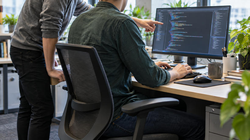 Synthetic editorial illustration. Two anonymous colleagues working together at a desk with a complete illuminated monitor directly in front of the seated engineer, who is using a keyboard; the colleague gestures toward softly rendered editor-like content.