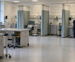Synthetic editorial illustration. A clinical treatment room contains curtained bays, reclining chairs and mobile equipment carts. A rear-facing nurse in navy scrubs attends to a volunteer's exposed arm at the far right while dark monitors stand nearby.