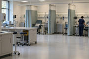 Synthetic editorial illustration. A clinical treatment room contains curtained bays, reclining chairs and mobile equipment carts. A rear-facing nurse in navy scrubs attends to a volunteer's exposed arm at the far right while dark monitors stand nearby.