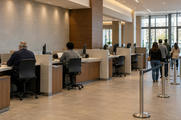 Synthetic editorial illustration. A bank branch interior. Two tellers viewed from behind sit at partitioned counters with edge-on monitors while a short queue of customers faces away beside the service area.