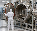 Synthetic editorial illustration. A white-suited technician viewed from behind holds open a large round stainless chamber door. Pipes, valves and vacuum fittings surround the open chamber in a bright clean industrial room.