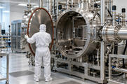 Synthetic editorial illustration. A white-suited technician viewed from behind holds open a large round stainless chamber door. Pipes, valves and vacuum fittings surround the open chamber in a bright clean industrial room.