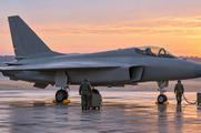 Synthetic editorial illustration. A plain grey single-tail jet stands on an airfield apron in warm dawn light. Two small crew members seen from behind attend unmarked wheeled equipment, with a low treeline beyond.