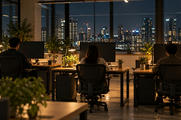 Synthetic editorial illustration. Three people sit with their backs to the viewer at separate desks with dark monitors, potted plants and warm task lamps. City lights fill the windows beyond the office.