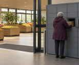 Synthetic editorial illustration. A supermarket entrance. A grey-haired shopper viewed from behind removes a cardboard box from a plain grey parcel locker, with loose fruit in wooden bins inside the glass doors.