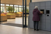 Synthetic editorial illustration. A supermarket entrance. A grey-haired shopper viewed from behind removes a cardboard box from a plain grey parcel locker, with loose fruit in wooden bins inside the glass doors.