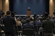 Synthetic editorial illustration. Rows of seated journalists and camera operators viewed from behind face a lectern in a formal press room. The distant speaker also faces away against a blank dark backdrop, with camera displays dark.