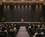 Synthetic editorial illustration. A large meeting hall contains an audience seated in rows facing an empty stage. Rear-facing attendees flank a central aisle leading to a plain wooden lectern against a dark blank backdrop.