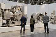 Synthetic editorial illustration. An equipment exhibition booth. Three visitors seen from behind look at large polished metal chambers and machinery on white platforms. Blank partition walls stand beneath the hall's exposed roof trusses.
