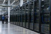 Synthetic editorial illustration. A server hall. Two rear-facing engineers stand beside long glass-fronted racks filled with equipment and small green, blue and amber lights, beneath overhead cable trays and bright strip lighting.
