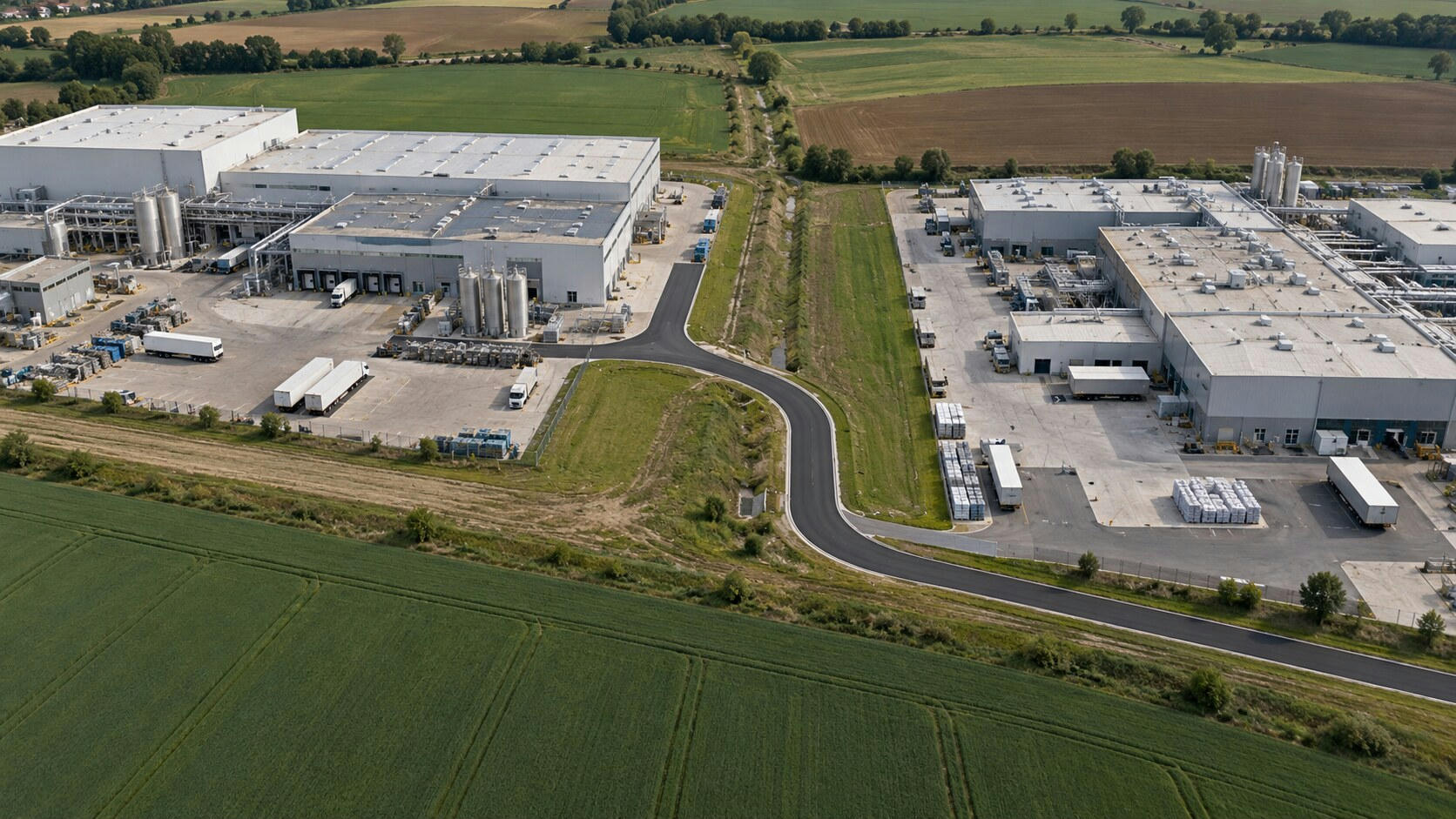 Synthetic editorial illustration. An industrial landscape viewed from above. Two neighbouring factory complexes with loading yards and metal tanks are linked by fresh dark asphalt through a grassy strip, surrounded by fields.