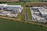 Synthetic editorial illustration. An industrial landscape viewed from above. Two neighbouring factory complexes with loading yards and metal tanks are linked by fresh dark asphalt through a grassy strip, surrounded by fields.
