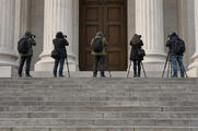 Synthetic editorial illustration. Broad courthouse steps lead to tall stone columns and closed wooden doors. Five rear-facing photographers wait on the upper landing with cameras, tripods and shoulder bags.