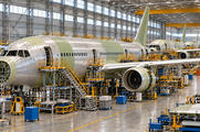 Synthetic editorial illustration. Several unpainted passenger-aircraft fuselages line a vast bright assembly hangar. Bare metal and pale primer surfaces sit among yellow access platforms, wheeled cabinets and overhead cranes.