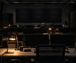 Synthetic editorial illustration. A network operations room contains rows of dark monitors and a wall of switched-off displays. Small desk lamps illuminate a distant rear-facing worker and equipment racks.