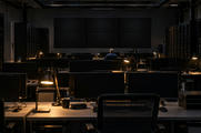Synthetic editorial illustration. A network operations room contains rows of dark monitors and a wall of switched-off displays. Small desk lamps illuminate a distant rear-facing worker and equipment racks.