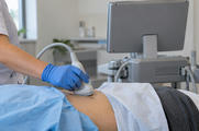 Synthetic editorial illustration. A blue-gloved hand holds an ultrasound probe against a draped patient's abdomen. The plain back of the monitor faces the camera beside unlabeled clinical supplies; no heads are visible.
