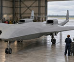 Synthetic editorial illustration. A broad unmarked aircraft with two upper engine inlets, twin tail booms and a straight joining tailplane stands in a hangar. Two engineers viewed from behind stand beside a tool chest under soft daylight.