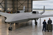 Synthetic editorial illustration. A broad unmarked aircraft with two upper engine inlets, twin tail booms and a straight joining tailplane stands in a hangar. Two engineers viewed from behind stand beside a tool chest under soft daylight.