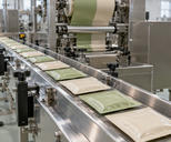 Synthetic editorial illustration. Plain cream and pale-green foil sachets travel along a stainless-steel conveyor beneath packing rollers. Bright windows light the clean industrial room.