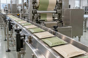 Synthetic editorial illustration. Plain cream and pale-green foil sachets travel along a stainless-steel conveyor beneath packing rollers. Bright windows light the clean industrial room.