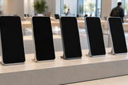 Synthetic editorial illustration. Five upright phones with entirely dark screens stand on plain metal supports along a pale showroom plinth. Daylight and softly blurred display tables fill the background.